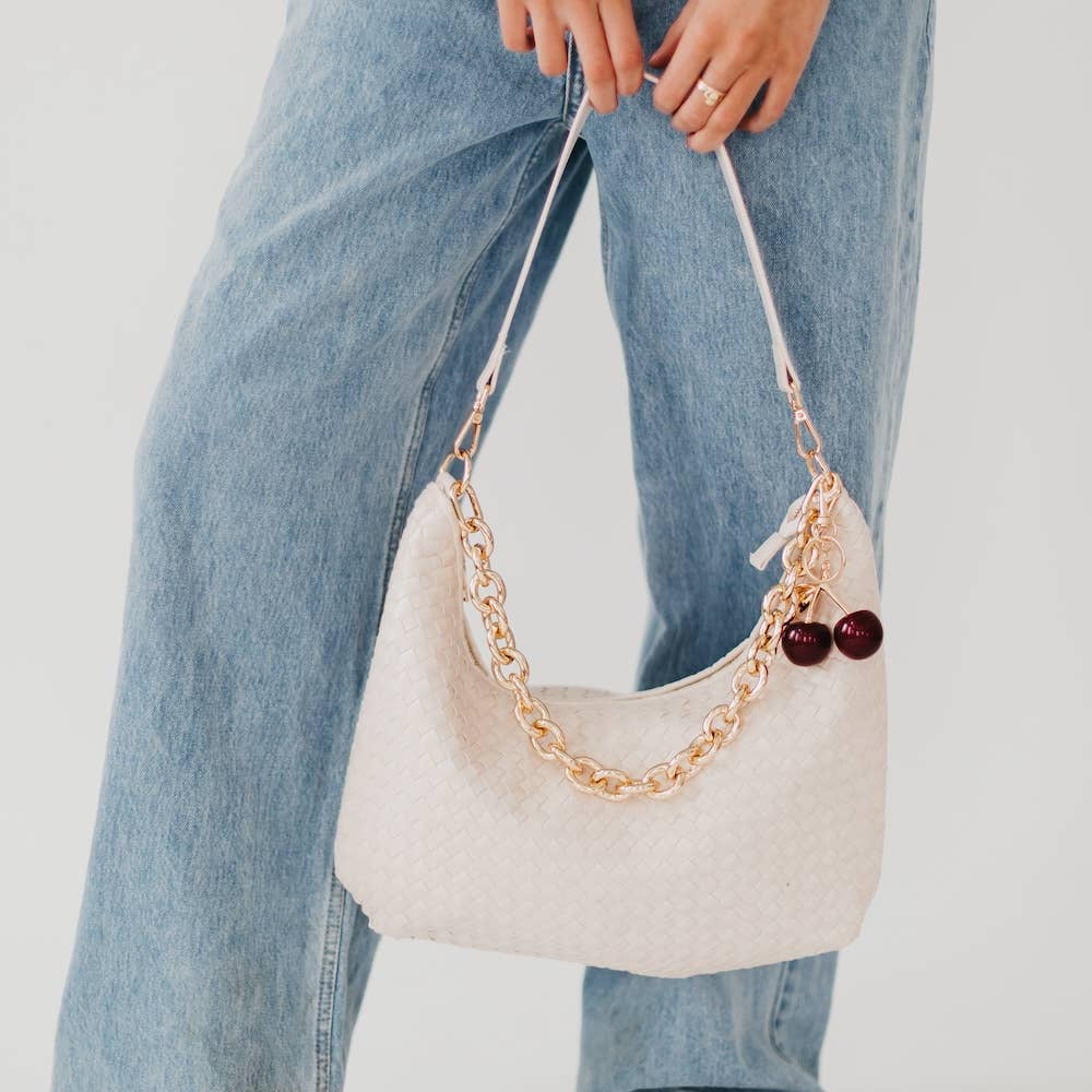 Beige handbag with gold chain and red beads held by a person wearing blue jeans on a light gray background