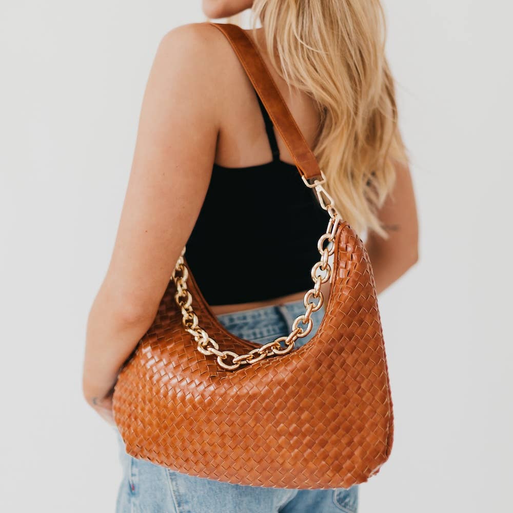 Person holding a brown woven handbag with a gold chain detail on a plain background